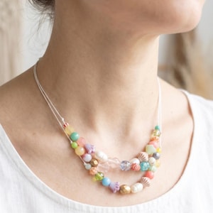 Op de afbeelding: Een dubbelstrengs ketting met een assortiment veelkleurige kralen, waaronder pastelkleurige bollen, bloemvormige bedels, witte parels en getextureerde metalen accenten geregen aan een dun wit koord, gedragen op een wit kledingstuk.