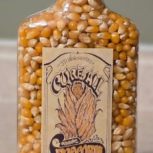 May include: A clear glass bottle filled with yellow popcorn kernels, topped with a black cap. The label reads "J.D. Winchesters Cure All Organic Popcorn" with directions for use. A vintage-style product.