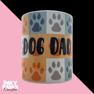 May include: A ceramic mug featuring a grid pattern of orange, blue, and grey paw prints. The text DOG DAD is printed in large, bold, dark blue letters across the centre of the mug, layered over the colourful paw print background design.