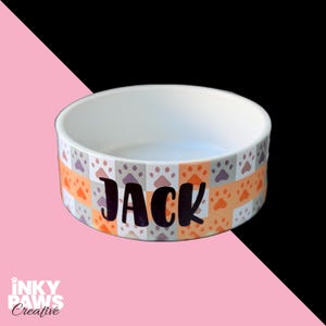 May include: A white ceramic pet bowl featuring a repeating checkered pattern of orange and grey paw prints around the exterior. The name JACK is printed in bold black lettering on the front. The interior of the bowl is plain white.