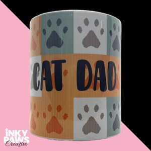 May include: A ceramic mug featuring a repeating pattern of paw prints in shades of blue, orange, and grey. A horizontal orange band across the centre displays the text CAT DAD in bold, dark blue letters. The design is set against a white background.