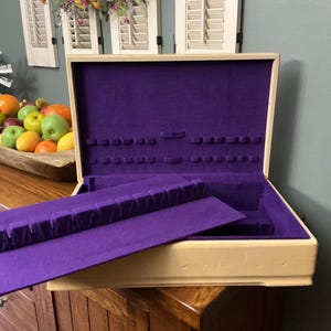 May include: Open wooden jewelry box with a cream-colored exterior and a vibrant purple velvet interior. The box is open, revealing compartments for storage. A removable tray sits inside. The box is on a wooden surface.
