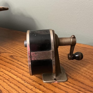 May include: Vintage pencil sharpener with a black and silver body and a bronze-colored handle. The sharpener is mounted on a metal base and sits on a wooden surface. The design is classic and functional.