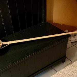 May include: A woven wicker basket with a long handle, likely a decorative or functional item. The basket is round with a circular bowl shape and a woven handle extending from the side. The handle is long and thin, and the basket is light brown.