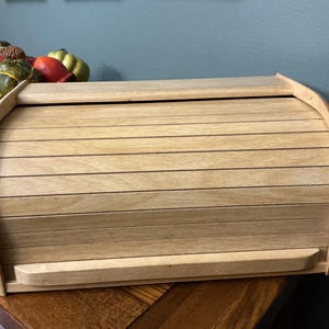 May include: A light-colored wooden bread box featuring a roll-top design with horizontal slats. The rectangular container has a curved top and a flat handle along the bottom edge of the sliding door, showcasing a natural wood grain finish throughout.