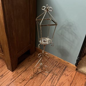 May include: A tall, silver-toned metal candle holder featuring a lantern-style top frame with a square opening, supported by a slender central rod and a decorative base composed of three curved, scroll-like legs that provide stability.