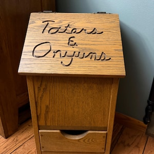 May include: A light brown wooden storage bin with a slanted hinged lid featuring the carved text Taters & Onyuns. The unit has a rectangular body with a lower drawer that includes a semi-circular cutout handle for easy access to stored items.