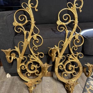 May include: A pair of gold-colored wall sconces featuring intricate scrollwork and swirling metal designs. Each sconce includes two candle holders with petal-shaped rims, extending from a tall, ornate vertical frame with decorative banding.