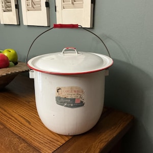 May include: White enamelware bucket with a red rim and handle. The bucket has a lid and a label with text. A black wire handle with a red grip arches over the top. The bucket sits on a wooden surface.