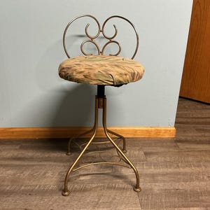 May include: A vintage vanity stool with a gold-colored metal frame and a round seat. The seat is upholstered in a patterned fabric with shades of green, brown, and tan. The back of the stool features a decorative heart-shaped design.