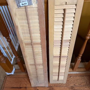 May include: Two wooden shutters, each with horizontal slats and a central control rod. One shutter is still in its packaging, with the text "Shutters" and "8x40" visible. The shutters are light brown and appear to be new.