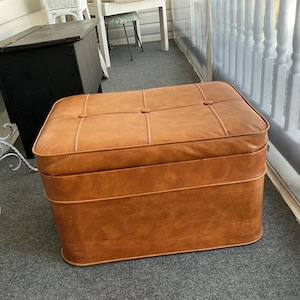 May include: A rectangular, brown leather ottoman with a tufted top and rounded edges. The ottoman has a lighter brown trim and is placed on a gray carpeted surface. The piece is a classic furniture item.