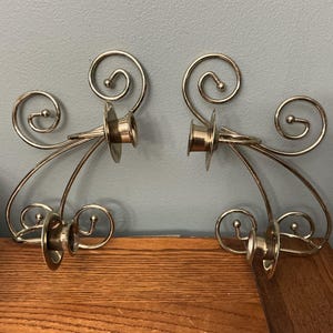 May include: A pair of silver-toned metal wall sconces featuring a curved, scrollwork design. Each sconce includes two circular candle holders with flat, round bases and decorative wire accents that terminate in small, rounded ball finials.