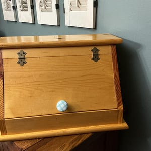 May include: A wooden bread box with a light brown finish. It features decorative metal hinges and a blue and white checkered knob. The box has a slanted front and a flat top.