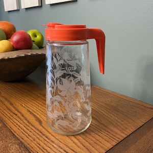 May include: Clear glass pitcher with an orange lid and handle. The pitcher features a floral design and a capacity marking of 1 qt. The pitcher is sitting on a wooden surface, with a wooden bowl of fruit in the background.