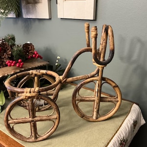 May include: A decorative bamboo tricycle planter with two wheels in the front and one in the back. The tricycle is made of light brown bamboo and has a handlebar and a seat for holding plants. The wheels have a cross-shaped design.