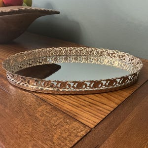 May include: An oval, gold-toned vanity tray with a mirrored surface. The tray's rim features a delicate, openwork design of leaves and scrolls. The tray sits on a wooden surface, with a wooden bowl holding fruit in the background.