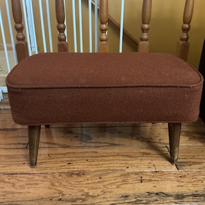 May include: A small, rectangular footstool with a dark reddish-brown upholstered top and four tapered wooden legs. The footstool is a vintage piece, likely from the mid-20th century, and is suitable for a living room or bedroom.