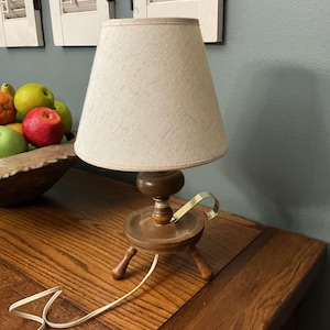 May include: A table lamp with a beige fabric shade and a dark brown wooden base. The base features a rounded central column, a circular platform, and three short legs. A curved gold-toned metal handle is attached to the side of the wooden base.