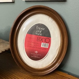 May include: An oval wall frame with a polished brown wood-tone finish. The frame contains a product insert labeled Empress Wall Frame 8x10, featuring a red and white design with a barcode and text detailing features like convex lens and mat for 5x7.