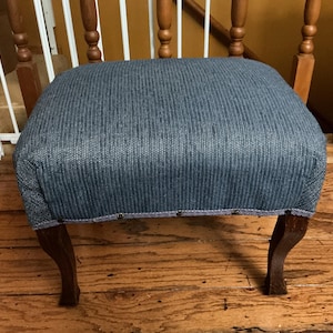 May include: A small footstool featuring a rectangular seat upholstered in a textured blue fabric with a subtle vertical stripe pattern. The base is supported by four dark brown wooden legs with a curved design, accented by metal nailhead trim.