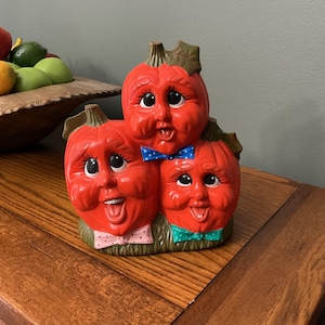 May include: A ceramic pumpkin figurine with three smiling faces, each wearing a bow tie. The pumpkins are red with green stems and leaves. The figurine is on a wooden surface.
