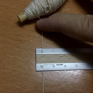 May include: A white thread is stretched across a wooden surface and measured with a silver ruler labeled "MIDORI MADE IN JAPAN". The ruler is 15 inches long.