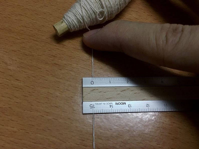 May include: A white thread is stretched across a wooden surface and measured with a silver ruler labeled "MIDORI MADE IN JAPAN". The ruler is 15 inches long.