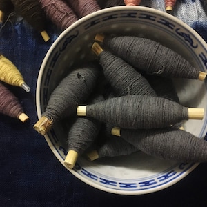 Puede incluir: Un bol lleno de carretes de hilo gris oscuro, cada uno con una clavija de madera. El bol es blanco con un patrón azul y blanco.