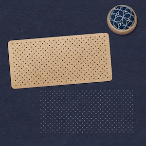 May include: A brown cardboard sheet with a grid of evenly spaced holes. The sheet is on a dark blue surface. A small, round, blue and white pincushion is in the upper right corner of the image.