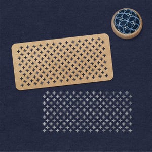 Könnte beinhalten: Ein braunes, hölzernes Kreuzstichmuster mit einem Gitter aus Pluszeichen. Das Muster befindet sich auf einem blauen Hintergrund und in der oberen rechten Ecke befindet sich ein kleines Nadelkissen mit einem weißen und blauen Design.