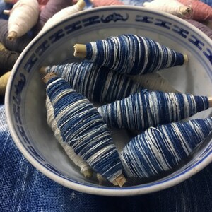 May include: A blue and white ceramic bowl filled with spools of indigo dyed thread. The spools are arranged in a circular pattern and have a natural wood center.