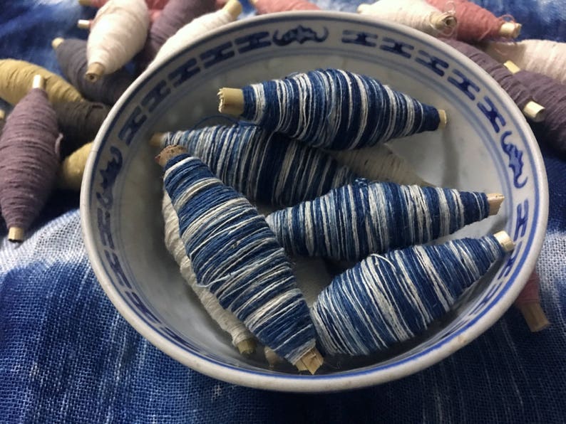 May include: A blue and white ceramic bowl filled with spools of indigo dyed thread. The spools are arranged in a circular pattern and have a natural wood center.