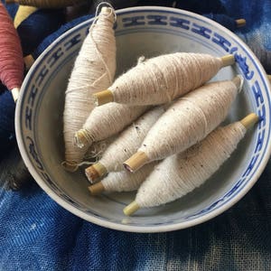 May include: A white ceramic bowl with a blue and white pattern holds several spools of natural colored yarn. The spools are made of wood and have a natural finish. The yarn is wound around the spools and is ready to be used for knitting or crocheting.