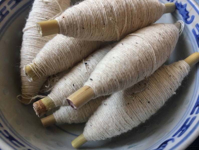 May include: A bowl filled with spools of white yarn wound around wooden dowels. The yarn is a natural off-white color and has a slightly textured surface.
