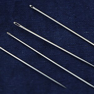 May include: Four silver sewing needles of varying lengths are arranged on a dark blue fabric background.