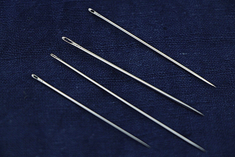 May include: Four silver sewing needles of varying lengths are arranged on a dark blue fabric background.