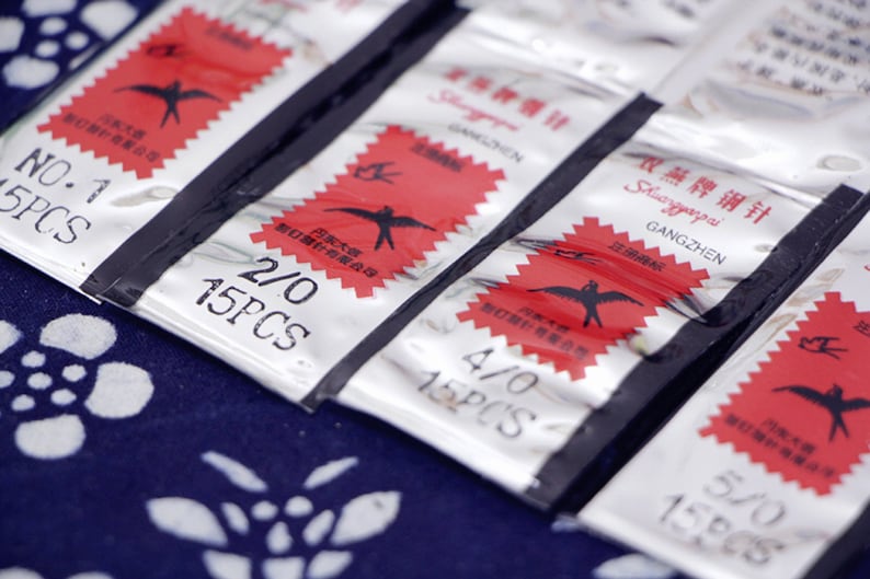 May include: Five silver packets of sewing needles with red labels. The labels feature a stylized bird design and the text "Shuangpai Gangzhen" in black. The packets are labeled with the sizes 1/0, 2/0, 4/0, and 5/0, and each packet contains 15 needles.