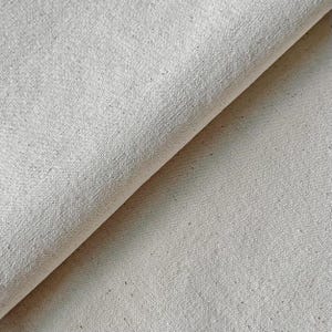 May include: Close-up of folded, off-white canvas fabric. The material has a textured surface with visible fibres and a slight diagonal fold, creating shadows and depth. The fabric appears to be a natural, unbleached cotton or linen.