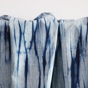 Op de afbeelding: Een close-up van een blauwe en witte tie-dye stof. De stof heeft een verticaal patroon van blauwe verf op een witte achtergrond.