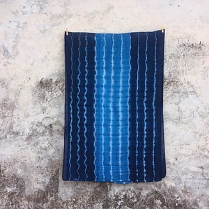 Op de afbeelding: Een blauwe en witte tie-dye stof die aan een muur hangt. De stof heeft een golvend patroon en is gemaakt van katoen.