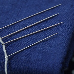 May include: Four silver needles are pinned into a blue fabric. A white thread is looped around the needles.