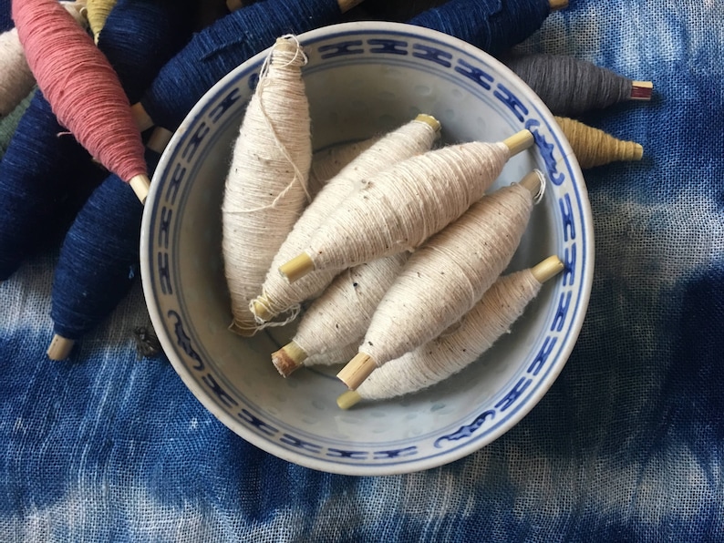 May include: A white ceramic bowl with a blue and white pattern holds several spools of natural colored yarn. The bowl is sitting on a blue and white fabric surface.