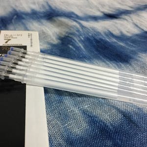 Könnte beinhalten: Eine Packung mit sieben transparenten, weißen, nachfüllbaren Tintenfüllern mit blauen Kappen. Die Füller sind gebündelt und liegen auf einem blau-weiß gemusterten Stoff.