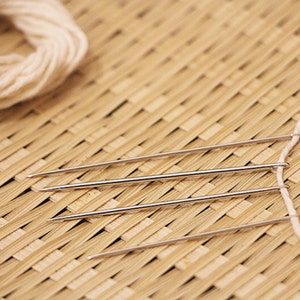 May include: Three silver needles are threaded with white yarn and laid out on a woven bamboo mat.
