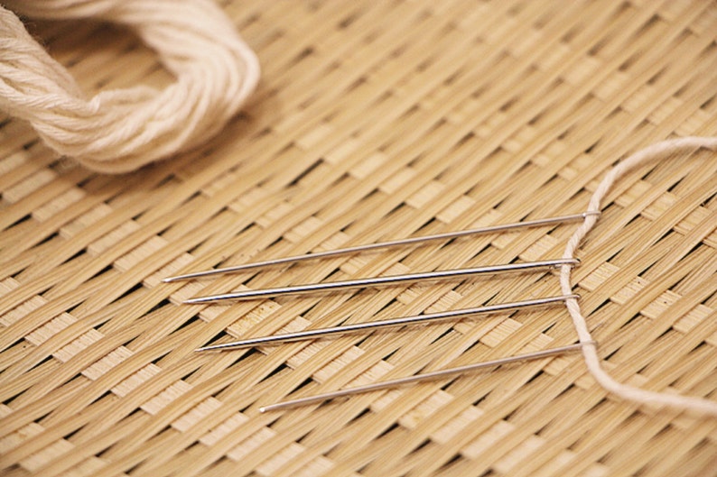 May include: Three silver needles are threaded with white yarn and laid out on a woven bamboo mat.
