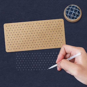 May include: A brown cardboard dot grid with holes for marking. A hand is using a white pen to transfer the dots onto a dark blue surface.