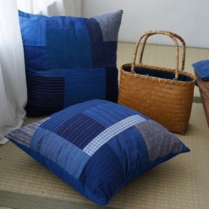 Puede incluir: Tres cojines azules de patchwork en varios tonos de índigo y mezclilla. Una bolsa de mimbre con asa está junto a los cojines. Los cojines están sobre una esterilla de tatami.