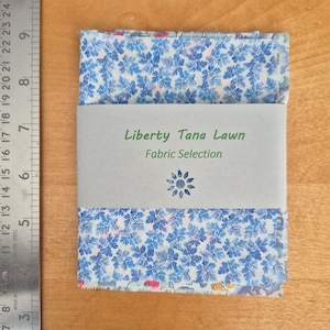 May include: A bundle of Liberty Tana Lawn fabric squares featuring a blue floral design. The fabric is wrapped with a grey band that reads "Liberty Tana Lawn Fabric Selection". A metal ruler is on the left.