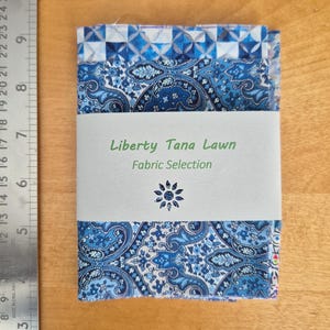 May include: A selection of Liberty Tana Lawn fabric, featuring a blue paisley design with a geometric border. The label reads "Liberty Tana Lawn Fabric Selection". The fabric is wrapped with a white band. The ruler shows the fabric is approximately 20 cm long.